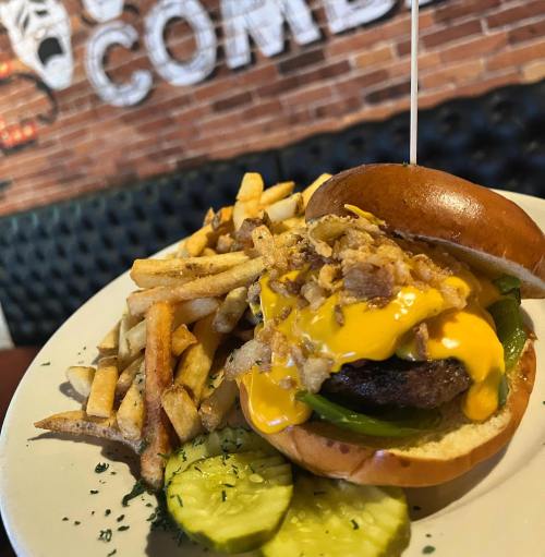 A close-up of a cheeseburger topped with fried onions and green peppers, served with fries and pickles on the side.