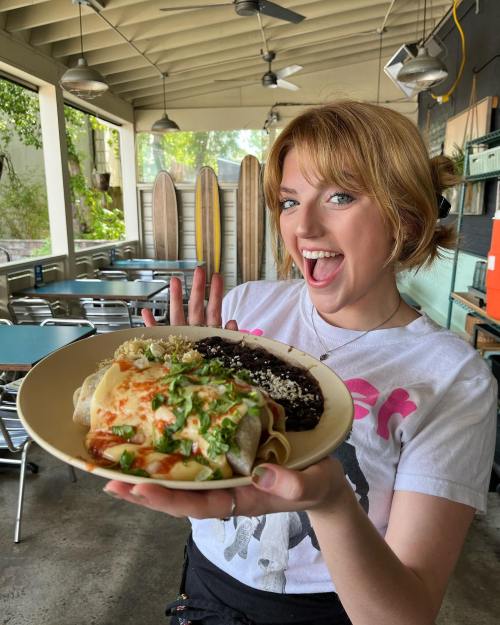 A smiling woman holds a plate of enchiladas with black beans and toppings in a casual dining setting.
