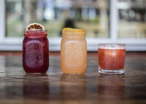 Three colorful drinks in jars and a glass, featuring a lemon slice and dried orange garnish, on a wooden table.