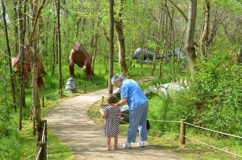 A child and an adult walk along a path in a forest, surrounded by large dinosaur sculptures among the greenery.