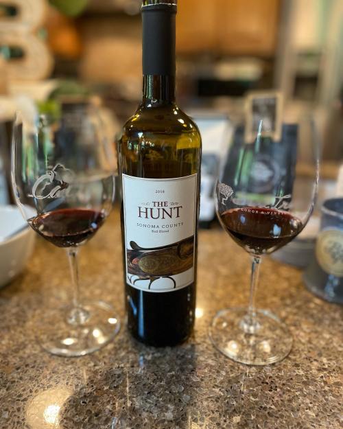 A bottle of red wine labeled "The Hunt" with two filled wine glasses on a countertop.
