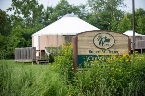 Sign for Robért W. Teater Conservation Area in front of a yurt, surrounded by greenery and wildflowers.