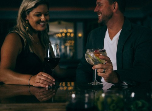 A couple enjoys drinks at a bar, smiling and engaging in conversation. The woman holds a glass of red wine, and the man has a cocktail.