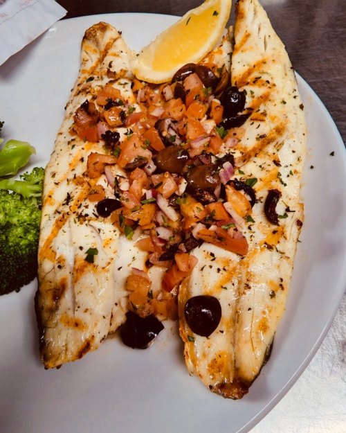Grilled fish fillets topped with diced tomatoes, olives, and herbs, served with a lemon wedge and broccoli on the side.