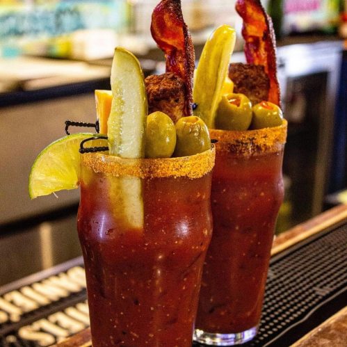 Two vibrant Bloody Mary cocktails garnished with bacon, pickles, olives, and a lime wedge, served in tall glasses.