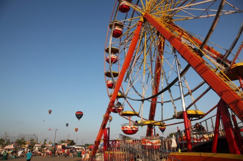 A colorful ferris wheel at a fair, with hot air balloons in the sky and people enjoying the festivities below.
