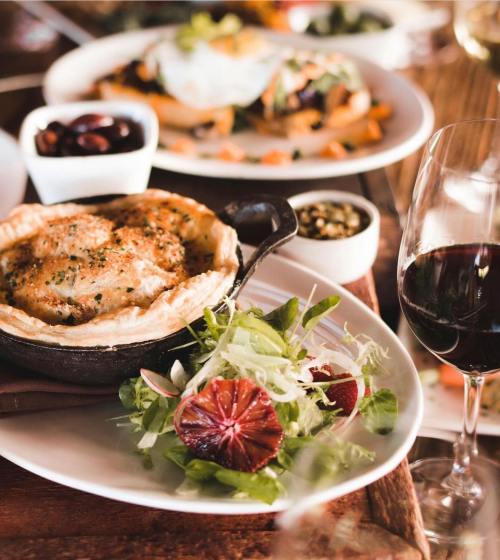 A savory pie in a skillet, fresh salad with blood orange, olives, and a glass of red wine on a rustic table.