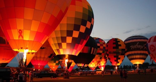 Colorful hot air balloons illuminated at dusk, with flames visible, creating a vibrant display against the evening sky.