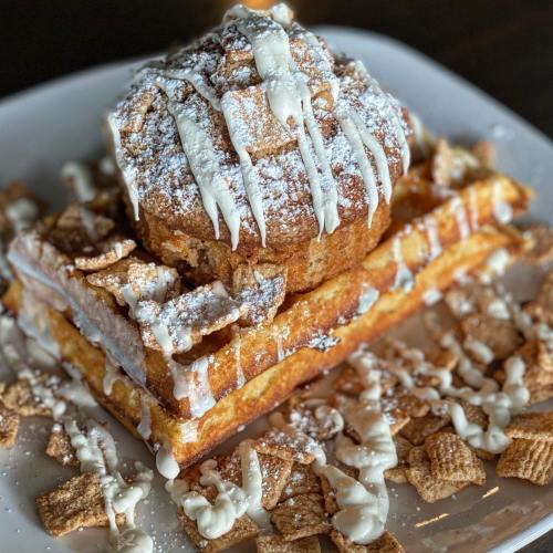 A stack of waffles topped with a cinnamon roll, drizzled with icing and surrounded by cereal pieces.