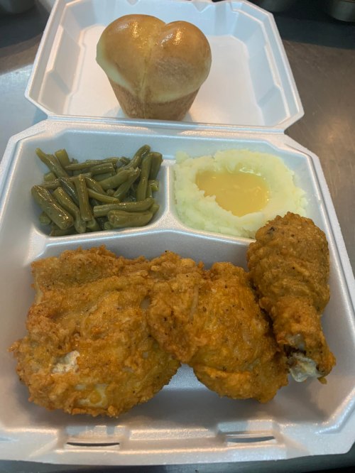 A takeout container with fried chicken, green beans, mashed potatoes with gravy, and a heart-shaped roll.