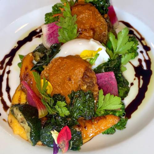 A colorful dish featuring crispy patties, greens, vibrant vegetables, and a poached egg, garnished with edible flowers.