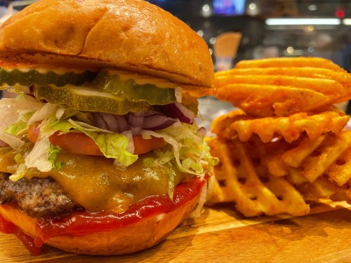 A close-up of a juicy burger with lettuce, tomato, pickles, and cheese, served with a side of crispy waffle fries.