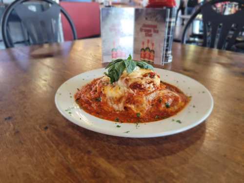 A plate of lasagna topped with cheese and basil, served in a red sauce, on a wooden table in a restaurant setting.