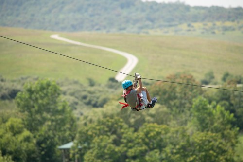 A person zip-lining over a green landscape with trees and a winding path in the background.