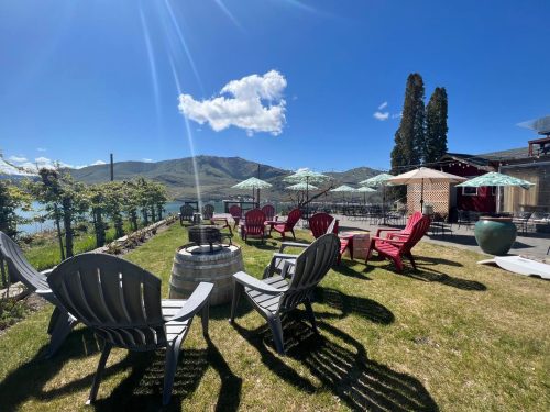 A scenic outdoor area with chairs, a fire pit, and umbrellas, overlooking mountains and a lake under a clear blue sky.