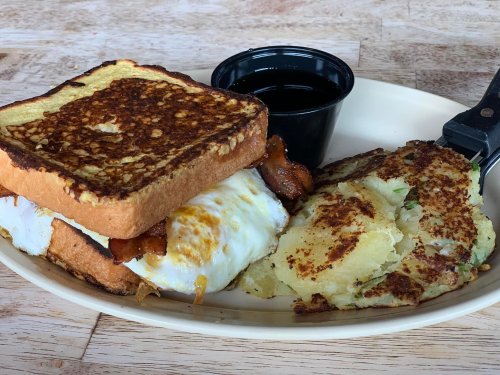 A plate with French toast, a fried egg, bacon, and crispy potato hash, served with a small cup of syrup.