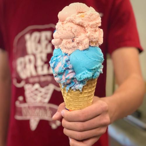 A person holds a tall ice cream cone with pink and blue scoops on top, wearing a red shirt with a logo.