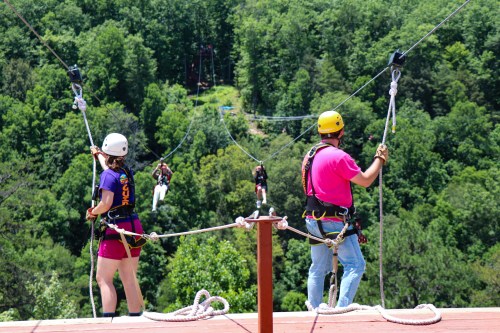 Two people in helmets and harnesses prepare to zip line over a lush green landscape, with others zipping in the background.