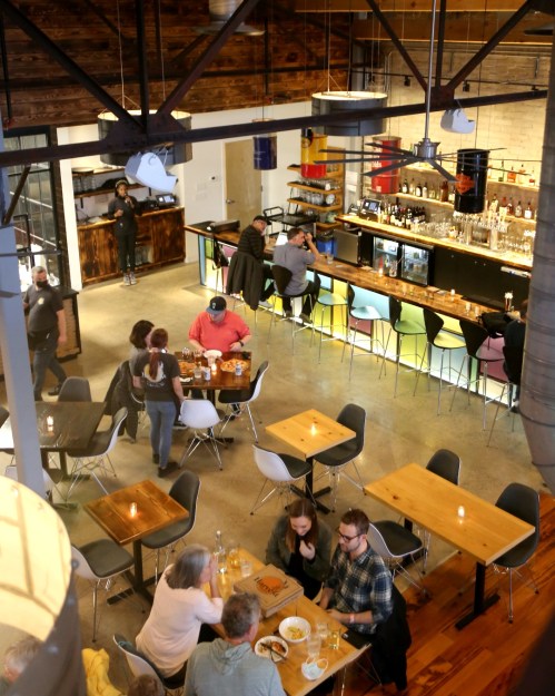 A modern restaurant interior with wooden tables, a bar, and patrons dining and socializing in a vibrant atmosphere.
