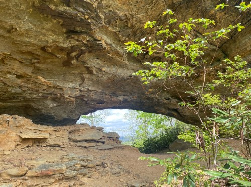 A natural rock arch with greenery and a view of the landscape beyond, under a cloudy sky.
