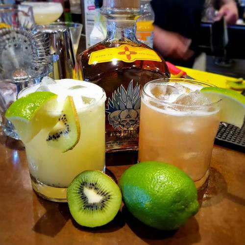 Two cocktails with lime and kiwi slices, a bottle of tequila in the background, and fresh limes on the bar.