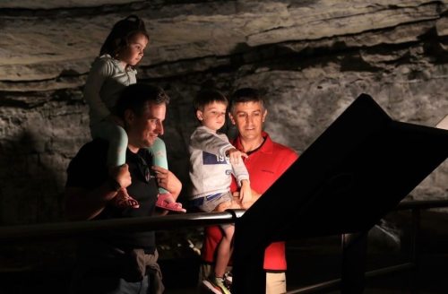 A man holds a child on his shoulders while another man and a girl look at a display in a dimly lit cave.