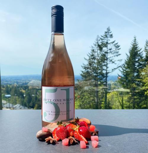 A bottle of Fifty One Weeks rosé wine surrounded by strawberries and candy, with a scenic view of trees and mountains in the background.