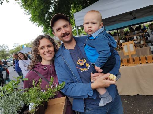 A smiling couple holds a child at a market, surrounded by plants and vendors in the background.