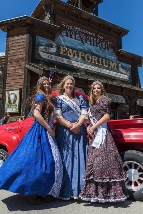 Three women in colorful dresses pose in front of a rustic building during a festive event in Winthrop.