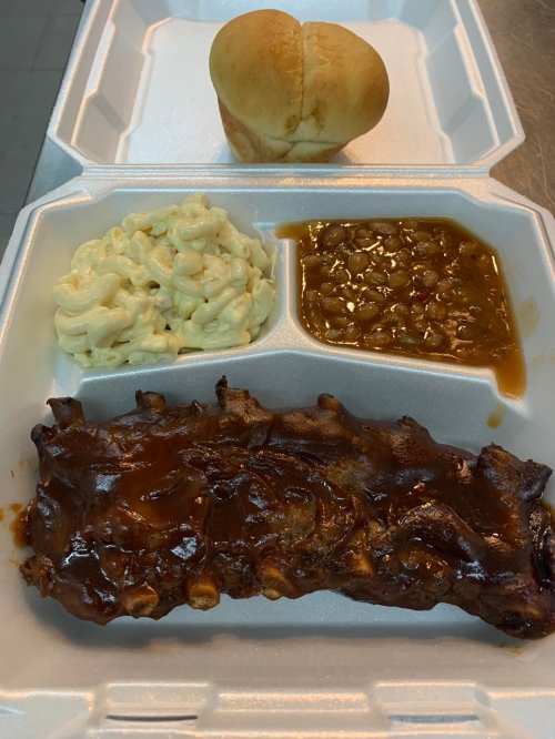 A takeout container with BBQ ribs, macaroni salad, baked beans, and a roll.