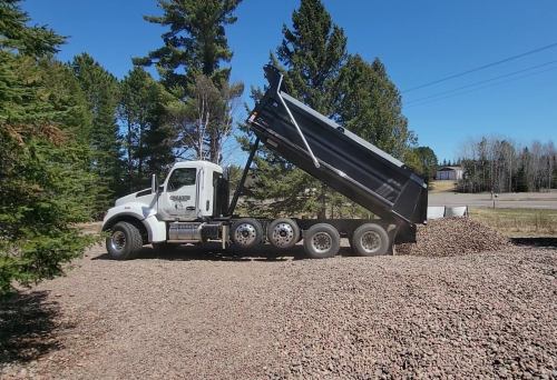A dump truck unloading gravel in a wooded area on a sunny day.