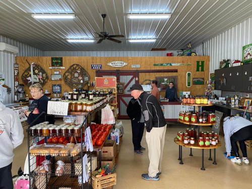 A busy store interior with customers browsing shelves of jars, produce, and local goods in a rustic setting.