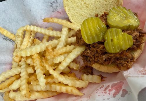 A pulled pork sandwich topped with pickles, served with a side of crinkle-cut fries on a paper-lined tray.