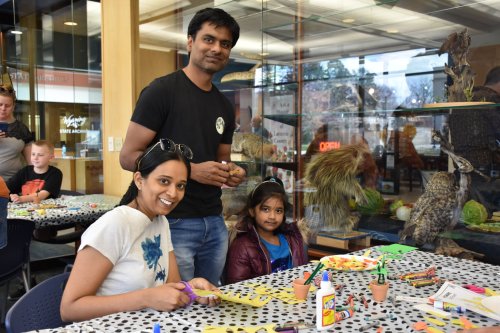A man and two children are crafting at a table filled with art supplies in a bright, cheerful environment.
