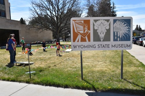 A sign for the Wyoming State Museum stands in front of a grassy area where children play and adults gather.