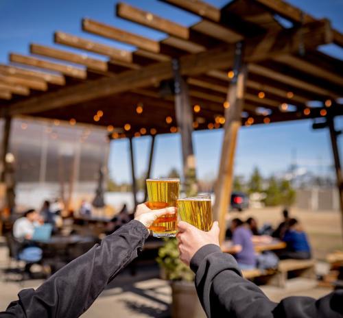 Two people clink glasses of beer outdoors under a wooden pergola, with a lively gathering in the background.
