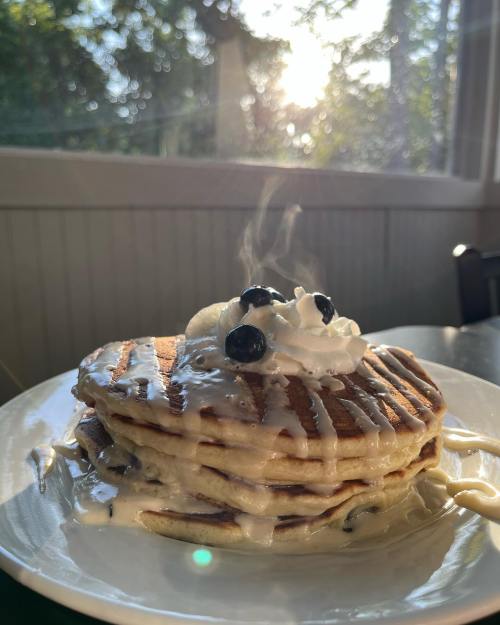 A stack of pancakes topped with whipped cream and blueberries, with steam rising, set against a sunlit background.