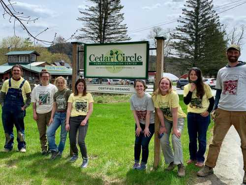 A group of eight people in matching shirts stands in front of the Cedar Circle Farm sign on a sunny day.