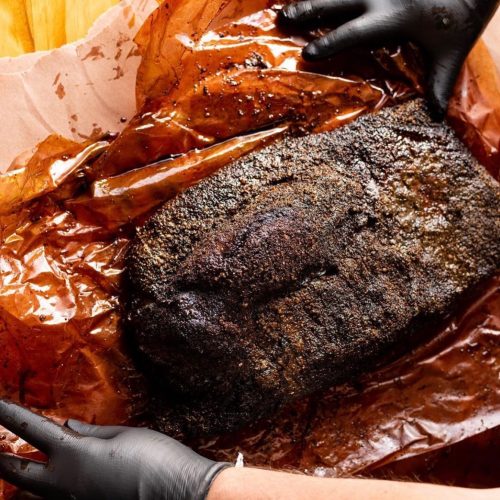 A person in black gloves holds a large, smoked brisket wrapped in brown butcher paper.