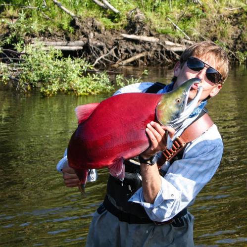 A person in sunglasses holds a large, vibrant red fish by the water, smiling proudly.