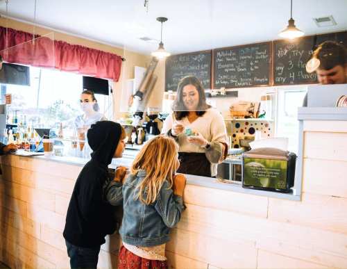 A barista serves two children at a café, with a chalkboard menu in the background and protective barriers in place.