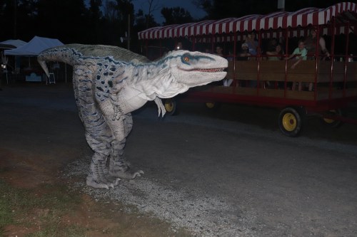 A dinosaur costume stands on a gravel path near a colorful train ride at an outdoor event.