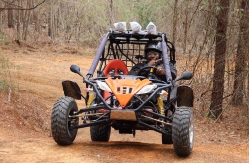 A person driving a go-kart on a dirt path surrounded by trees, wearing a helmet and holding the steering wheel.
