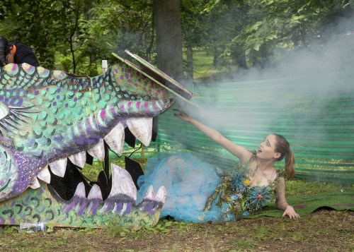 A woman in a blue dress reaches toward a colorful dragon sculpture, surrounded by greenery and smoke.