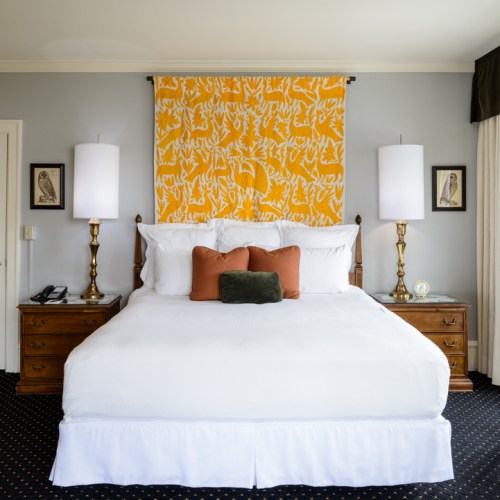 A cozy hotel room featuring a white bed, decorative pillows, and a vibrant yellow wall hanging. Elegant lamps on nightstands.
