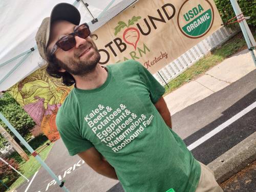 A smiling man in a green shirt with vegetable names stands under a tent at a farmers market, promoting Rootbound Farm.