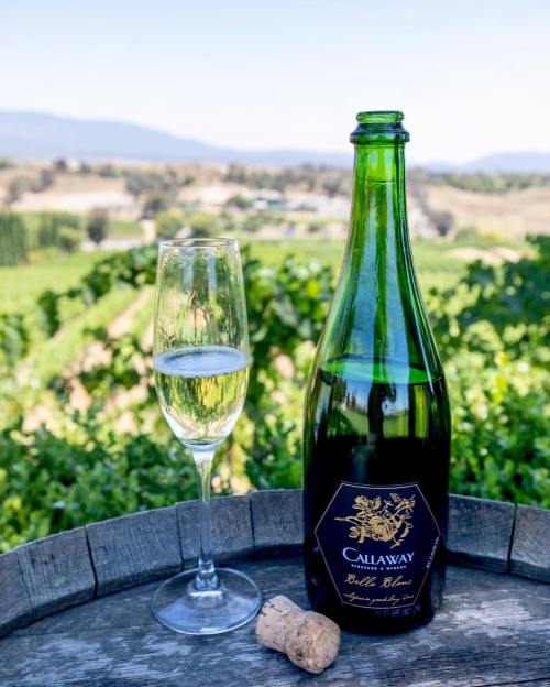 A bottle of Callaway sparkling wine with a glass, set against a vineyard backdrop and a cork on a wooden table.
