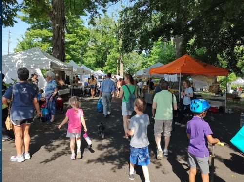 A bustling outdoor market with people walking among vendor tents, trees, and a sunny atmosphere.