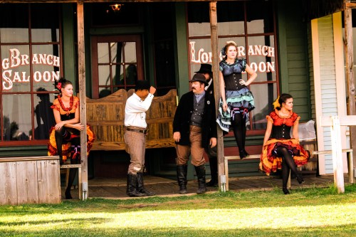 A group of performers in period costumes outside a saloon, with two women seated and three others interacting.