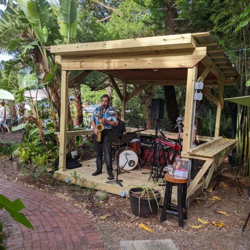 A musician plays saxophone on a wooden stage surrounded by greenery, with drums and equipment nearby.